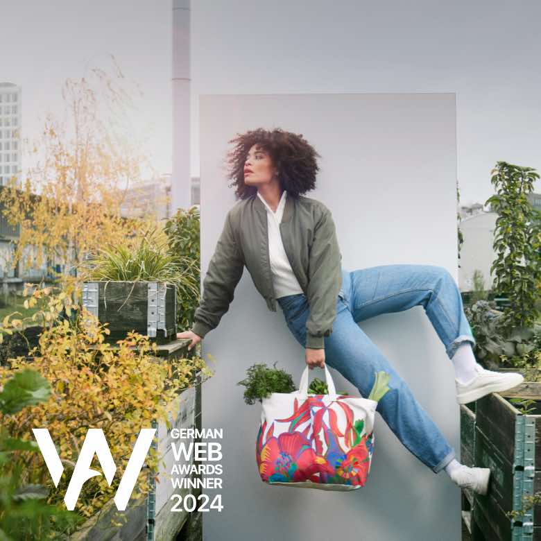 Kampagne für 17Ziele. Entwicklung einer limitierten Fashion Kollektion mit Designer Dawid Tomaszewski, Foto- und Videoshooting, Pre- und Postproduktion, Entwicklung von Social-Media-Assets, 18/1 und City-Light-Postern, Webdesign und Postkarten. Auszeichnung: German Web Award Winner 2024. Bild: Eine junge Frau in Bomberjacke und Jeans hält einen bunt gemusterten Shopper und posiert dynamisch an einer weißen Wand vor urbanem Hintergrund mit Pflanzen. Text: TRAG ES IN DIE WELT.