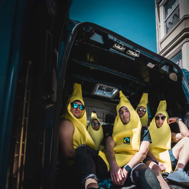 Social-Media-Kampagne für Fairtrade Deutschland. Konzeption, Fotoshooting mit Mitarbeitenden, Postproduktion, Schnitt und Gestaltung von Reels und Captions für Social Media, Kampagnenplanung, Kampagnenauswertung, Community Management. Bild: Fünf Personen sitzen gedrängt im Laderaum eines Transporters; alle tragen leuchtend gelbe Bananenkostüme und Sonnenbrillen und blicken cool in die Kamera. Text: VOLL IM FAIRTRADE-FIEBER.